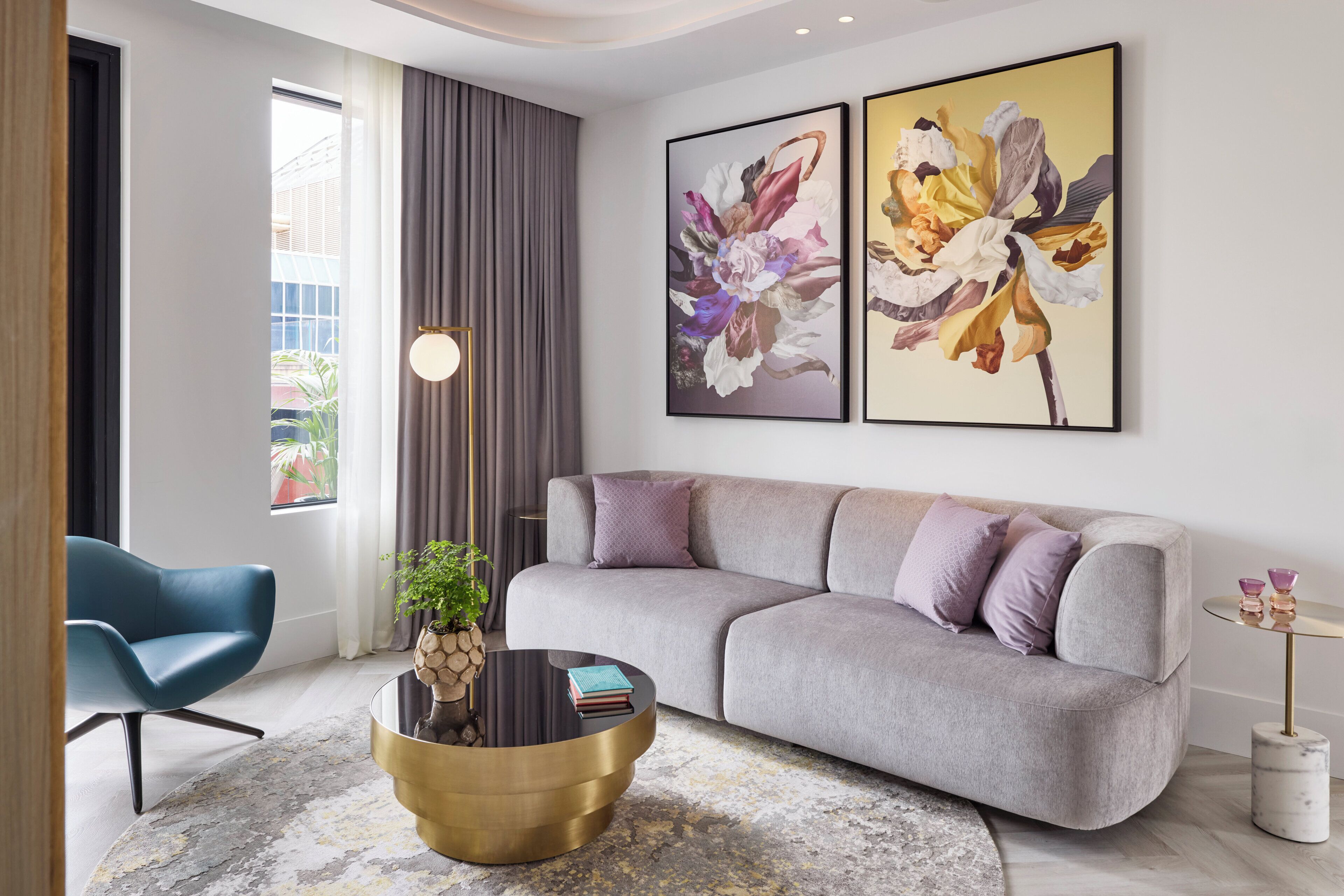 Una sala de estar moderna con un sofá gris, un sillón azul, una mesa de centro redonda y dos cuadros con motivos florales en la pared.