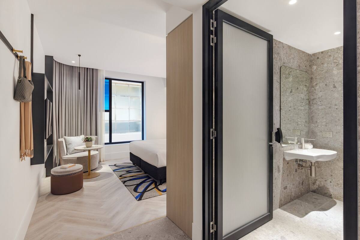 Ein modernes Hotelzimmer mit Bett, einer Sitzecke mit Tisch und Stühlen, einem Badezimmer mit Waschbecken und einem großen Fenster, das für natürliches Licht sorgt.