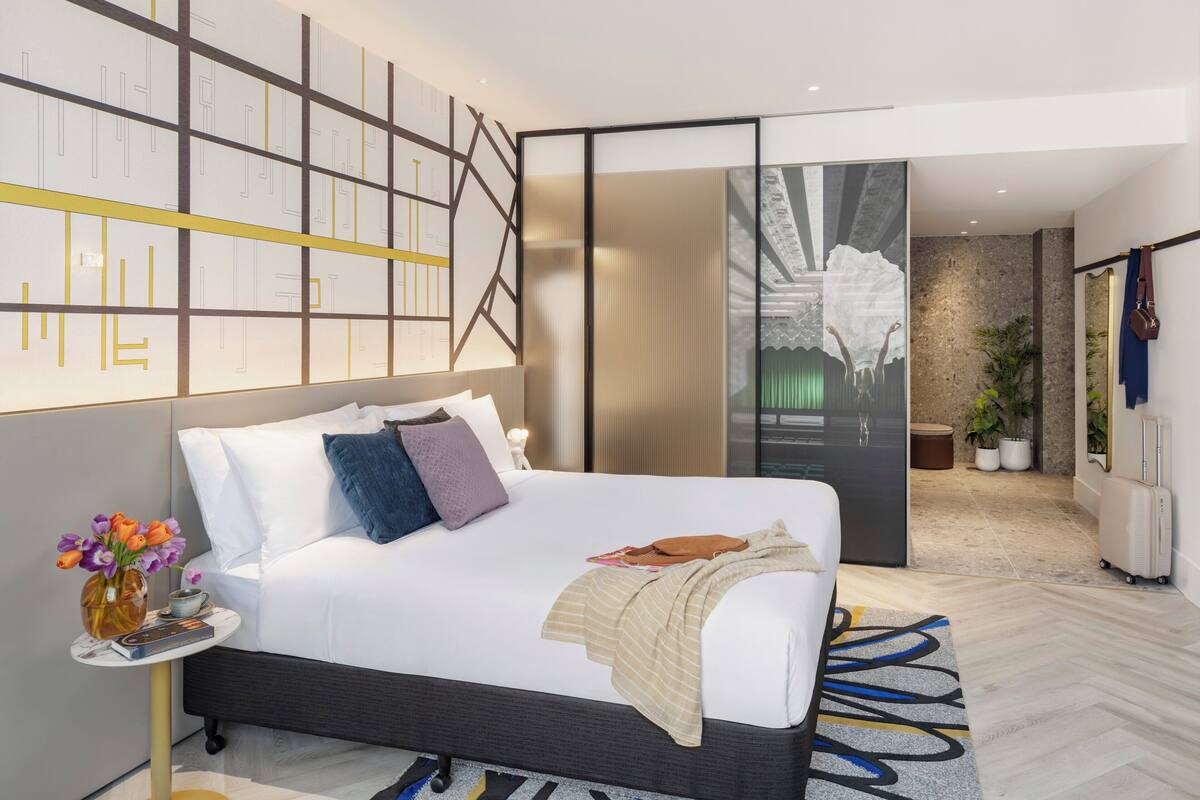 Ein modernes Hotelzimmer mit einem großen Bett, einem Nachttisch mit Blumen in einer Vase, einer Wand mit geometrischen Mustern und Blick auf die Stadt durch eine Glastür.