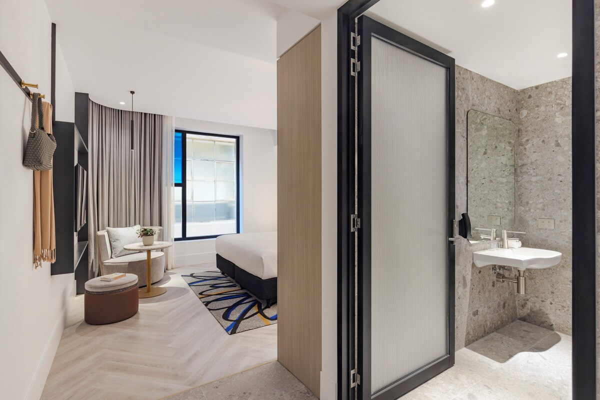 Ein modernes Hotelzimmer mit Bett, einer Sitzecke mit Tisch und Stühlen, einem Badezimmer mit Waschbecken und einem großen Fenster, das für natürliches Licht sorgt.