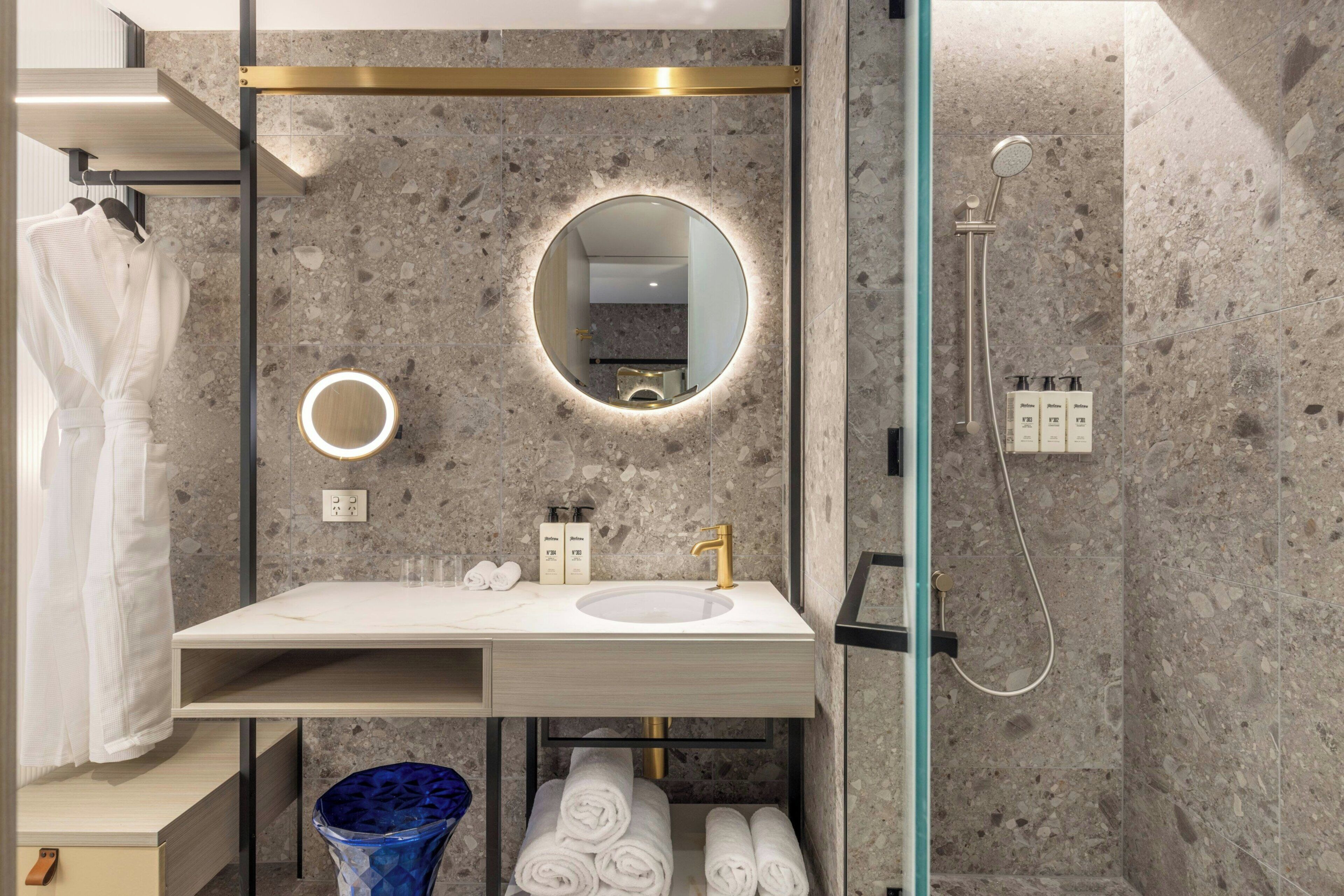 Um banheiro moderno com bancada de mármore, espelho redondo, torneira dourada e chuveiro com divisória de vidro.