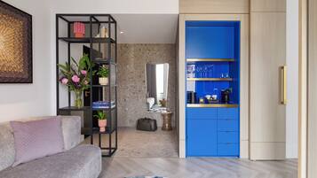 Una sala moderna con un sofá gris, un estante negro con libros y flores, un armario azul con utensilios de cocina y una alfombra estampada.