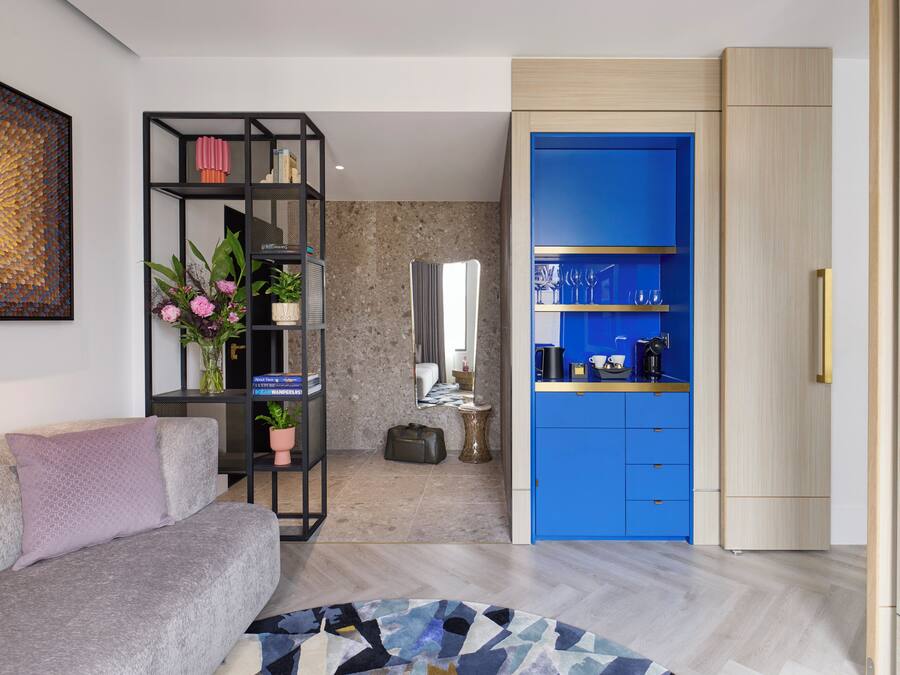 Una sala moderna con un sofá gris, un estante negro con libros y flores, un armario azul con utensilios de cocina y una alfombra estampada.