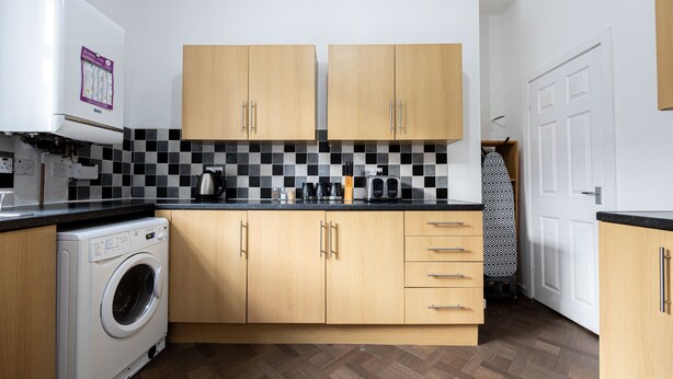 A compact kitchen with a washing machine, wooden cabinets, a black and white checkered backsplash, and a patterned door. - Barnes Street Terrace (Accrington, Lancashire, United Kingdom)
