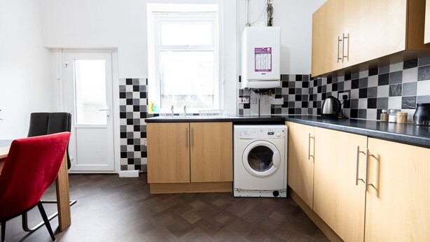 A compact kitchen with a washing machine, wooden cabinets, and a checkered backsplash. - Barnes Street Terrace (Accrington, Lancashire, United Kingdom)