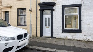 A white car parked in front of a building with a white and black facade, a black door with the number 77, and a barred window. - Barnes Street Terrace (Accrington, Lancashire, United Kingdom)