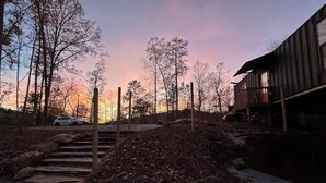 Uma cabana de madeira com uma escadaria de pedra que leva a um deck, rodeada por árvores e um céu ao pôr do sol.