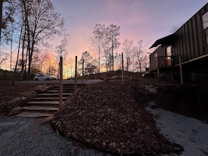 Una cabaña de madera con una escalera de piedra que lleva a una terraza, rodeada de árboles y un cielo al atardecer.
