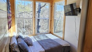 A bedroom with a large window, a bed, a TV mounted on the wall, and a view of trees outside. - Eagle Nest Cabin B at Walterfalls – Upstate SC (Six Mile)