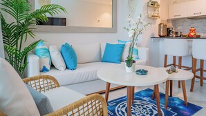 Una sala de estar moderna con un sofá blanco, cojines azules, una mesa de centro redonda y una zona de cocina con armarios blancos y una encimera.