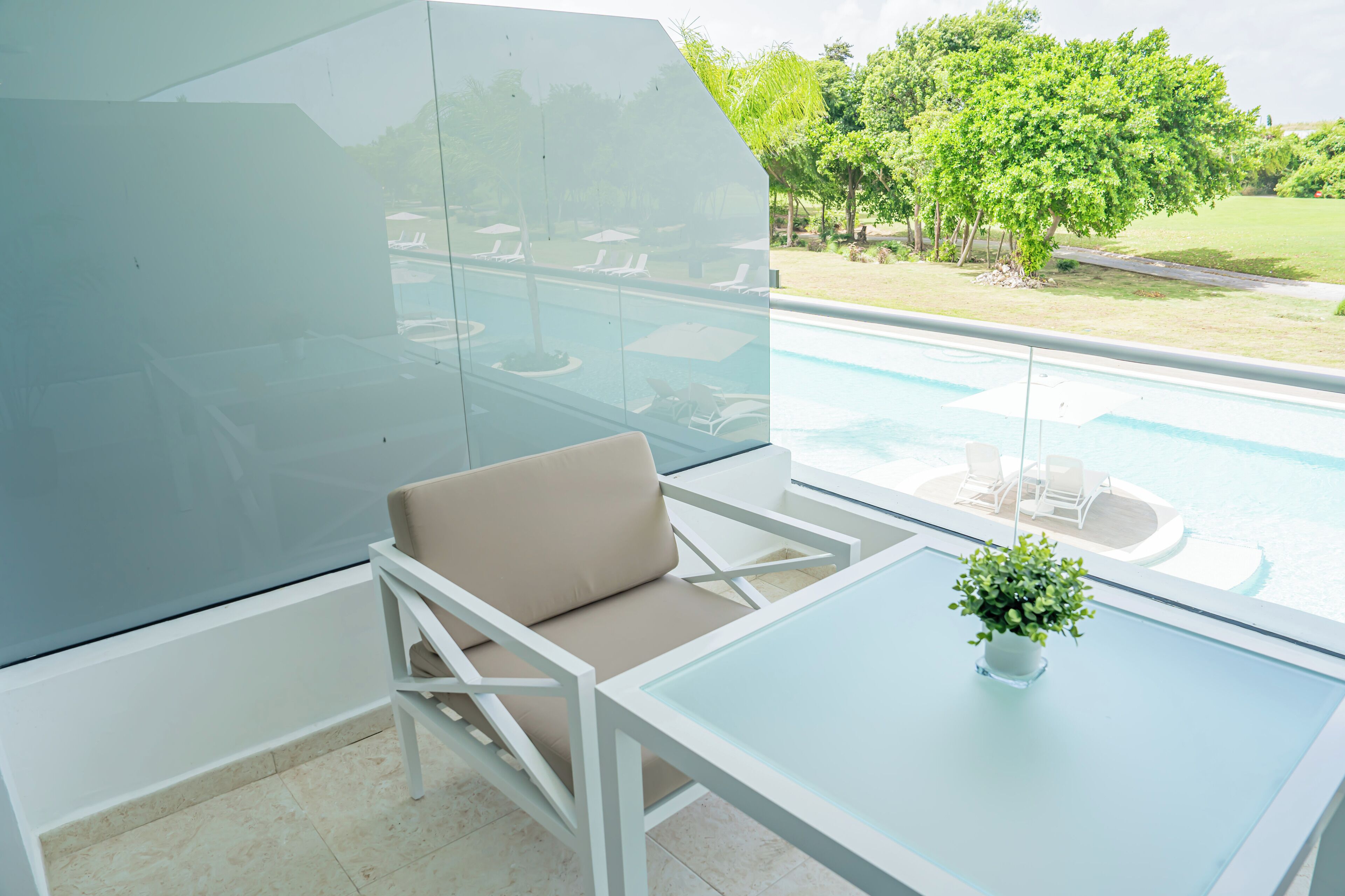A modern outdoor seating area with a glass table and a cushioned chair, overlooking a swimming pool and a landscaped garden.