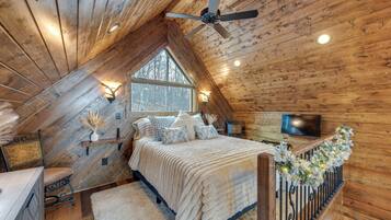 A cozy attic bedroom with a wooden ceiling, a large window, a bed with a white comforter, a TV, and a decorative railing.