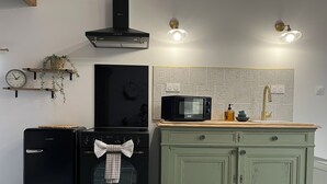 Ett kök med ett svart kylskåp, en mikrovågsugn och en spis. Där finns ett grönt skåp med lådor, en klocka på väggen och en växt på en hylla.