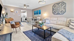 Ein modernes Wohnzimmer mit Sofa, Couchtisch und Sesseln. Ein Essbereich mit Tisch und Stühlen ist ebenfalls vorhanden. Im Hintergrund ist eine Küche zu sehen.