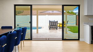 A modern kitchen with a large island, a dining area with blue chairs, and a sliding glass door leading to a backyard with a pool and patio furniture. - Sexy Boutique Estate in Fun Size - Swank Chill Vibe - Mill Ave - Spring Training (Tempe)