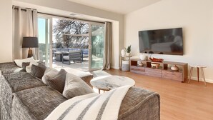 Een moderne woonkamer met een grote bank, een salontafel en een flatscreen televisie aan de muur. Er is een glazen schuifdeur die leidt naar een buitenpatio.