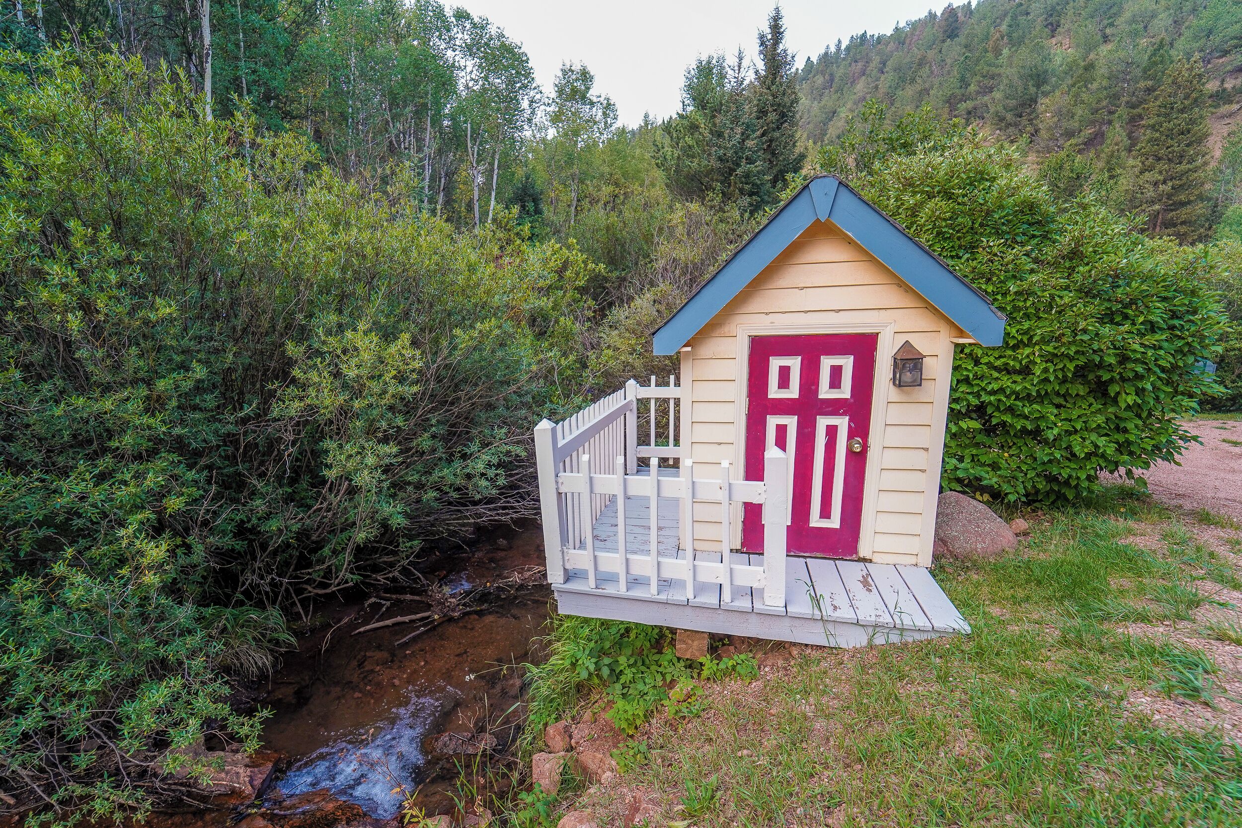 Uma pequena cabana de madeira com porta vermelha e varanda branca, rodeada por vegetação densa e um riacho.