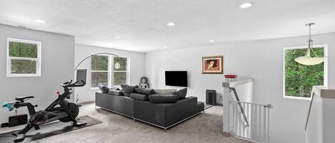 A spacious living area with a gray sofa, a standing exercise bike, and a staircase leading to an upper level.