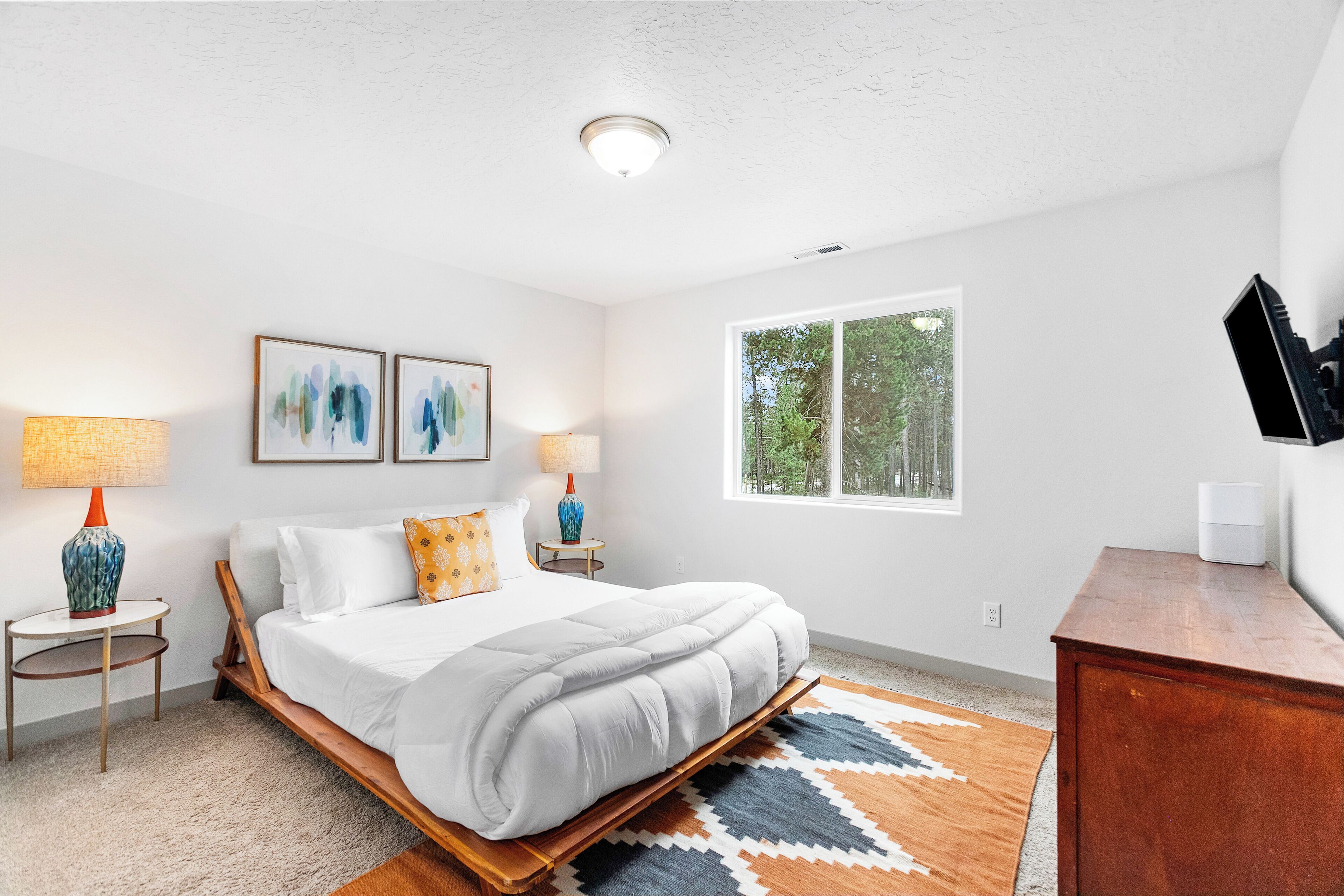 A bedroom with a bed, two bedside tables with lamps, a window, and a TV on a wooden dresser.
