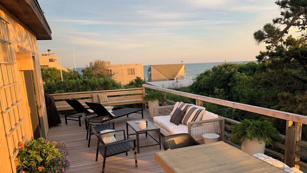 A wooden deck with outdoor furniture, including a sofa and chairs, overlooking a body of water. - Fire Island Pines Oasis w/4 King Bdrms, Heated Pool, Hot Tub, Ocean & Bay Views (Sayville)