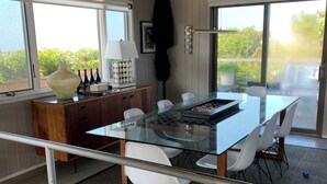 A modern dining area with a glass table, white chairs, and a wooden cabinet with a wine rack. - Fire Island Pines Oasis w/4 King Bdrms, Heated Pool, Hot Tub, Ocean & Bay Views (Sayville)