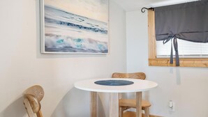 A room with a round wooden table, two chairs, a framed painting of a wave, and a window with a curtain.