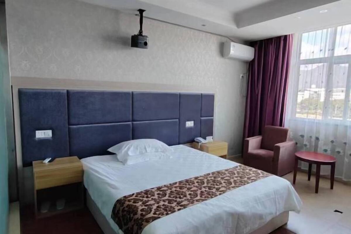 A hotel room with a bed, a chair, a small table, a nightstand, and a ceiling-mounted projector.