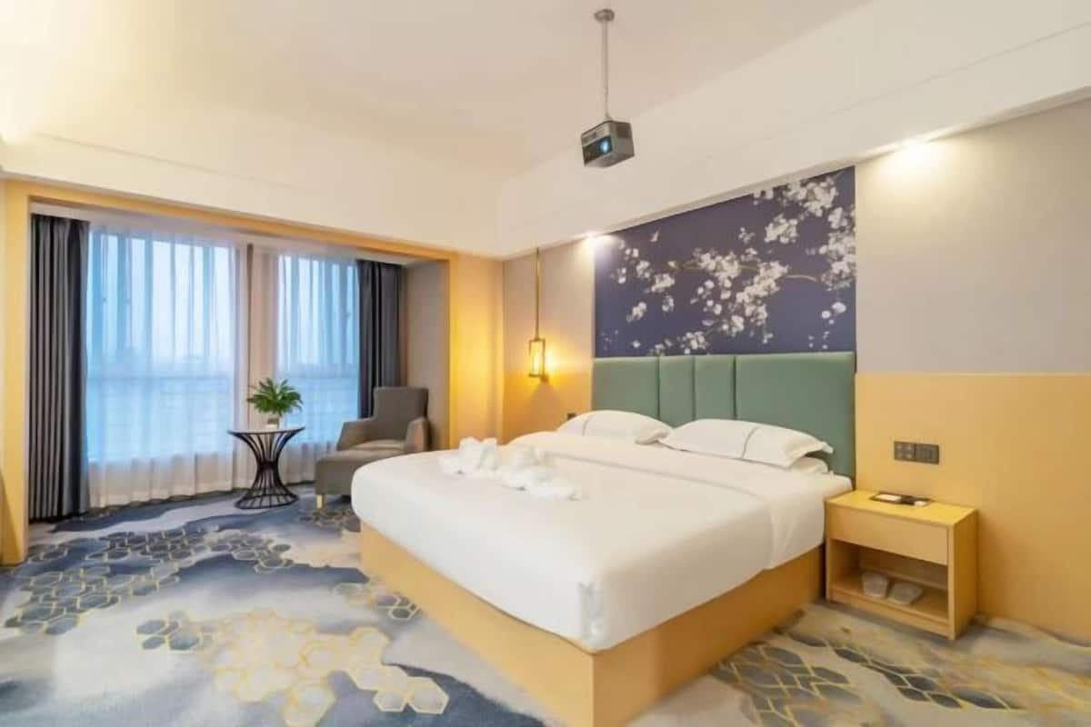 A hotel room with a bed, bedside table, lamp, and a view of the outdoors through large windows.