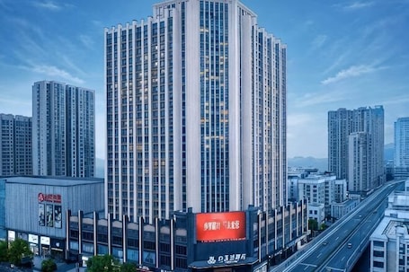 A high-rise building with a prominent red sign at the base, surrounded by other tall buildings and a highway.. Magnotel Hotel (Yichang CBD Shopping Center)
