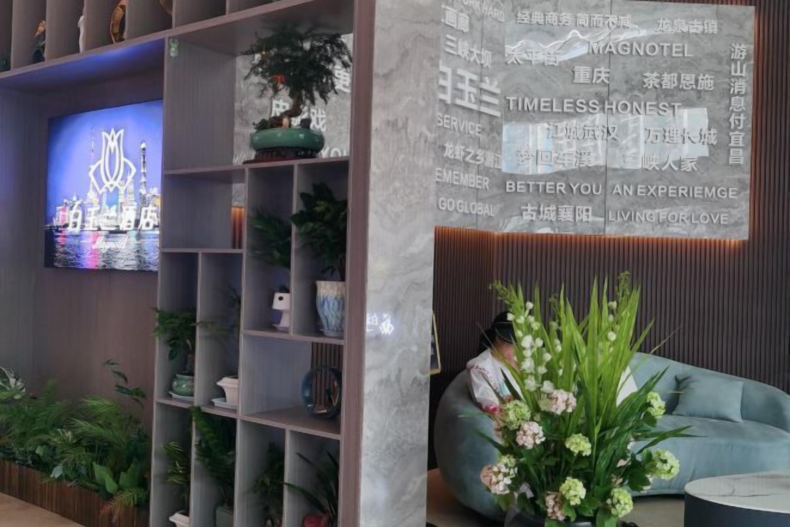 A modern hotel lobby with a large TV screen displaying a logo, a reception desk, and a seating area with a floral arrangement.