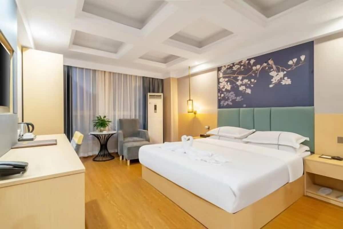 A hotel room with a bed, a desk, a chair, a small table with a plant, and a wall with a floral mural.