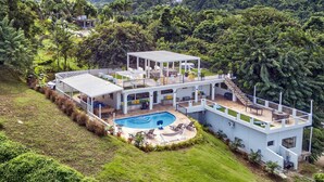 Ein modernes, mehrgeschossiges Haus mit Pool, umgeben von üppiger Vegetation und Bergen.