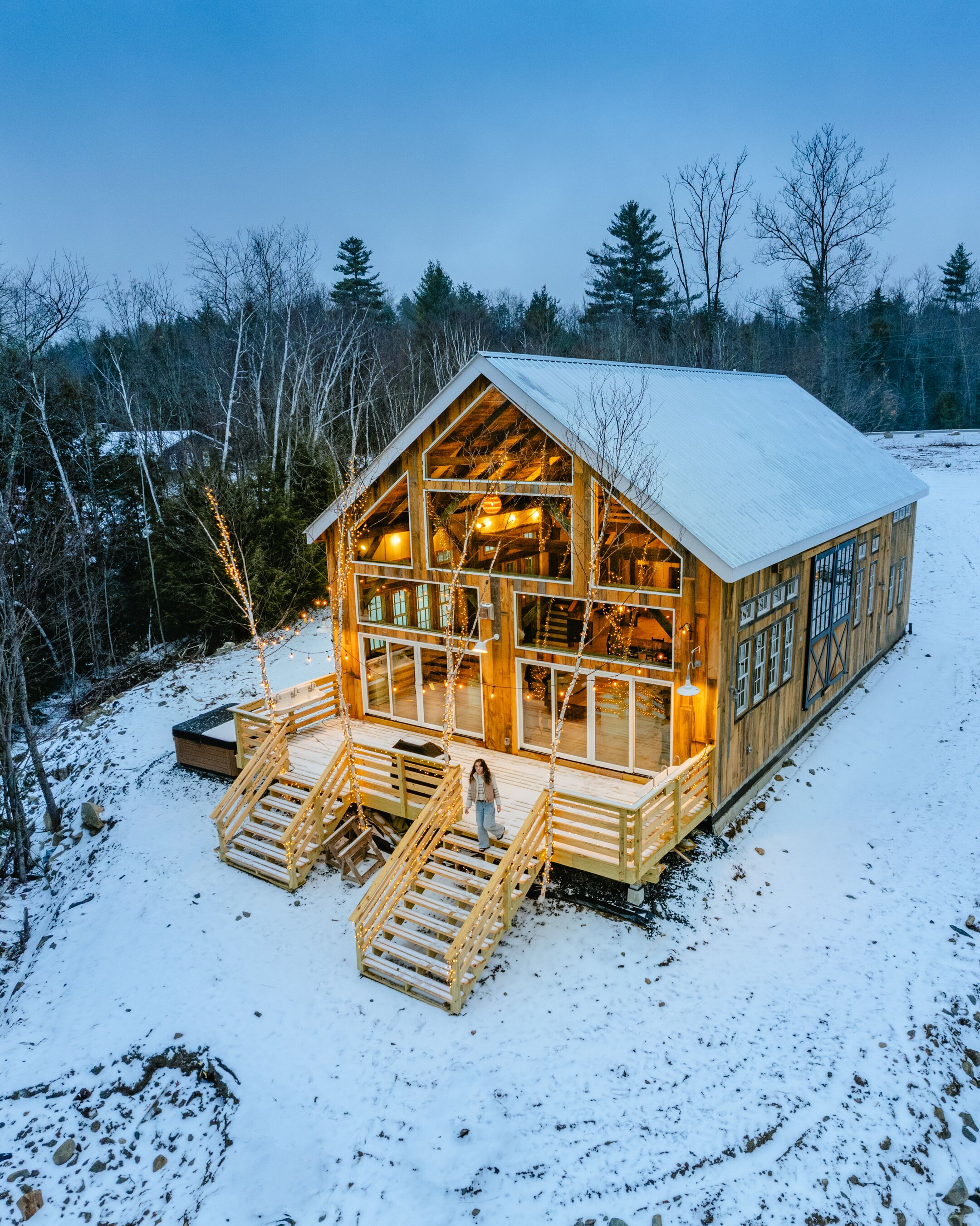 Une cabane en bois dotée d’une terrasse et d’un escalier recouverts de neige, entourée d’arbres.
