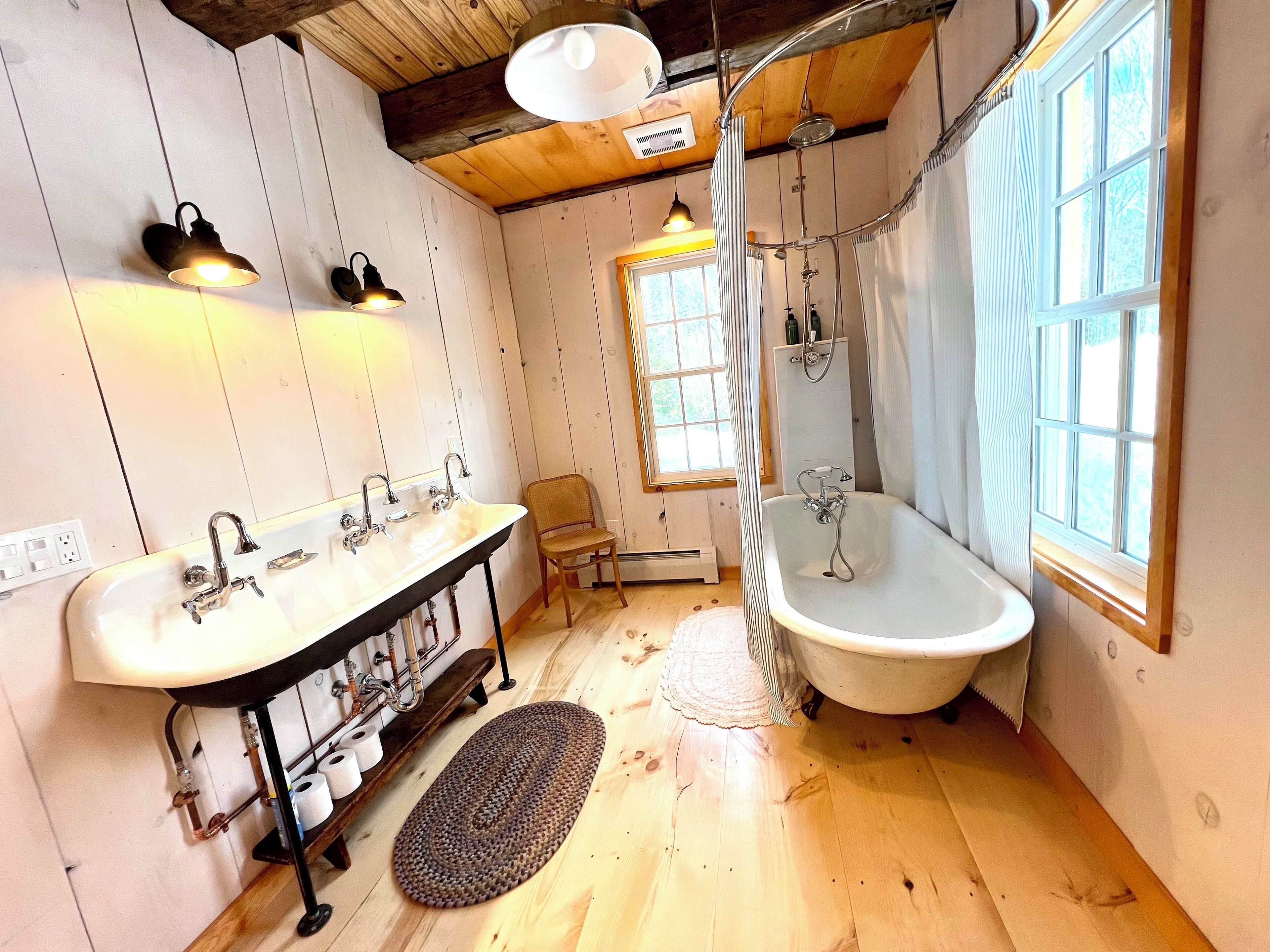 A rustic bathroom with a wooden floor, a large bathtub, a double sink, and a window providing natural light.