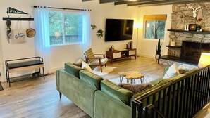 A spacious living room with a green sofa, a wooden coffee table, and a fireplace. There is a television mounted on the wall and a stone fireplace surround. - Charming 4-bedroom house with AC and WiFi in awesome Crestline (Crestline)