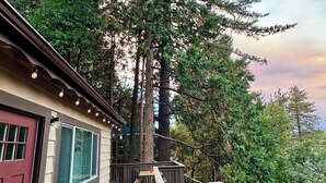 A wooden deck with a red door, a window with a view of tall trees, and a small table and chair. - Charming 4-bedroom house with AC and WiFi in awesome Crestline (Crestline)