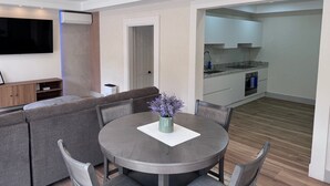 Uma sala de estar moderna com um sofá cinzento, uma mesa de jantar redonda com cadeiras, uma televisão de tela plana e uma cozinha com armários brancos e forno embutido.