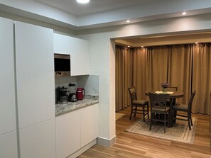 A compact kitchen with white cabinets, a coffee maker, and a blender. There is a dining area with a table and chairs, and a window with curtains.