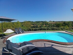 Une piscine entourée d’une terrasse en bois, agrémentée de chaises longues et d’un parasol, et offrant une vue sur un paysage verdoyant.