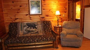 Une pièce chaleureuse avec un mur en bois, un canapé agrémenté d’un coussin aux motifs naturels, un fauteuil, une petite table avec une lampe et un tableau encadré accroché au mur.
