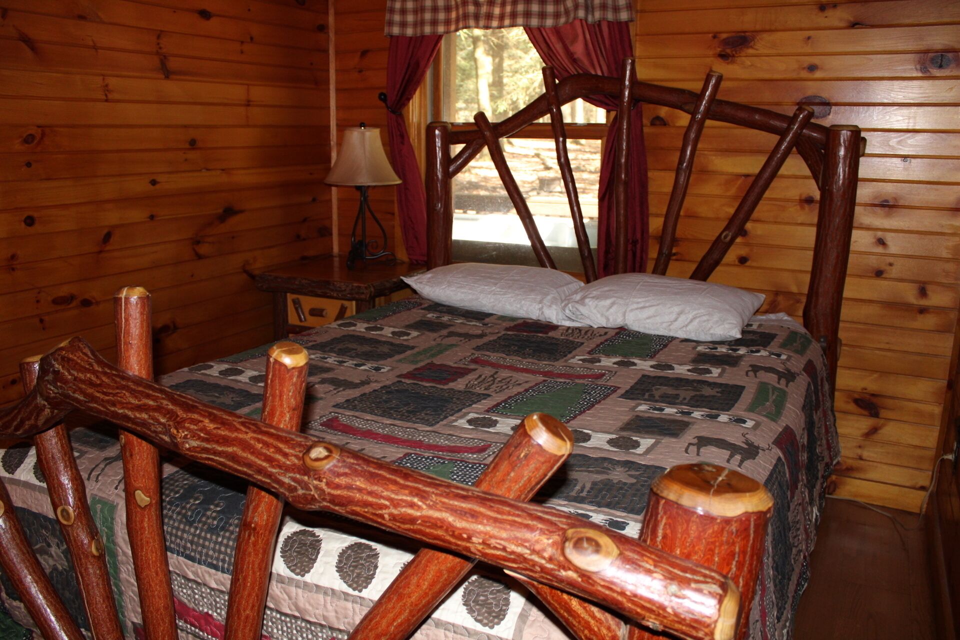 A wooden cabin bedroom with a bed, a nightstand, a lamp, and a window with curtains.