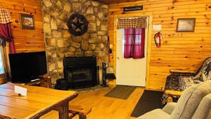 L’intérieur chaleureux d’un chalet avec une cheminée en pierre, un téléviseur, une table en bois et un fauteuil confortable.