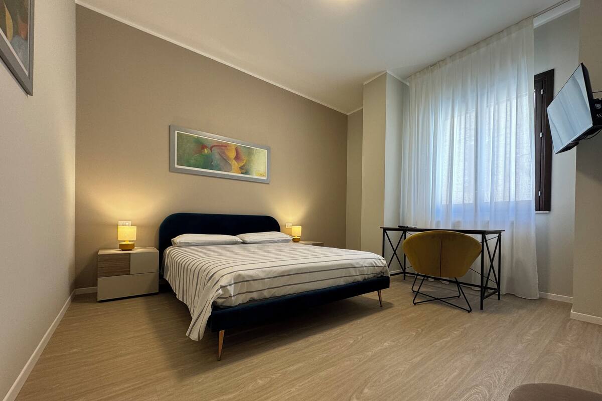 A modern hotel room with a bed, bedside lamps, a desk with a chair, a painting on the wall, and a TV mounted on the wall.