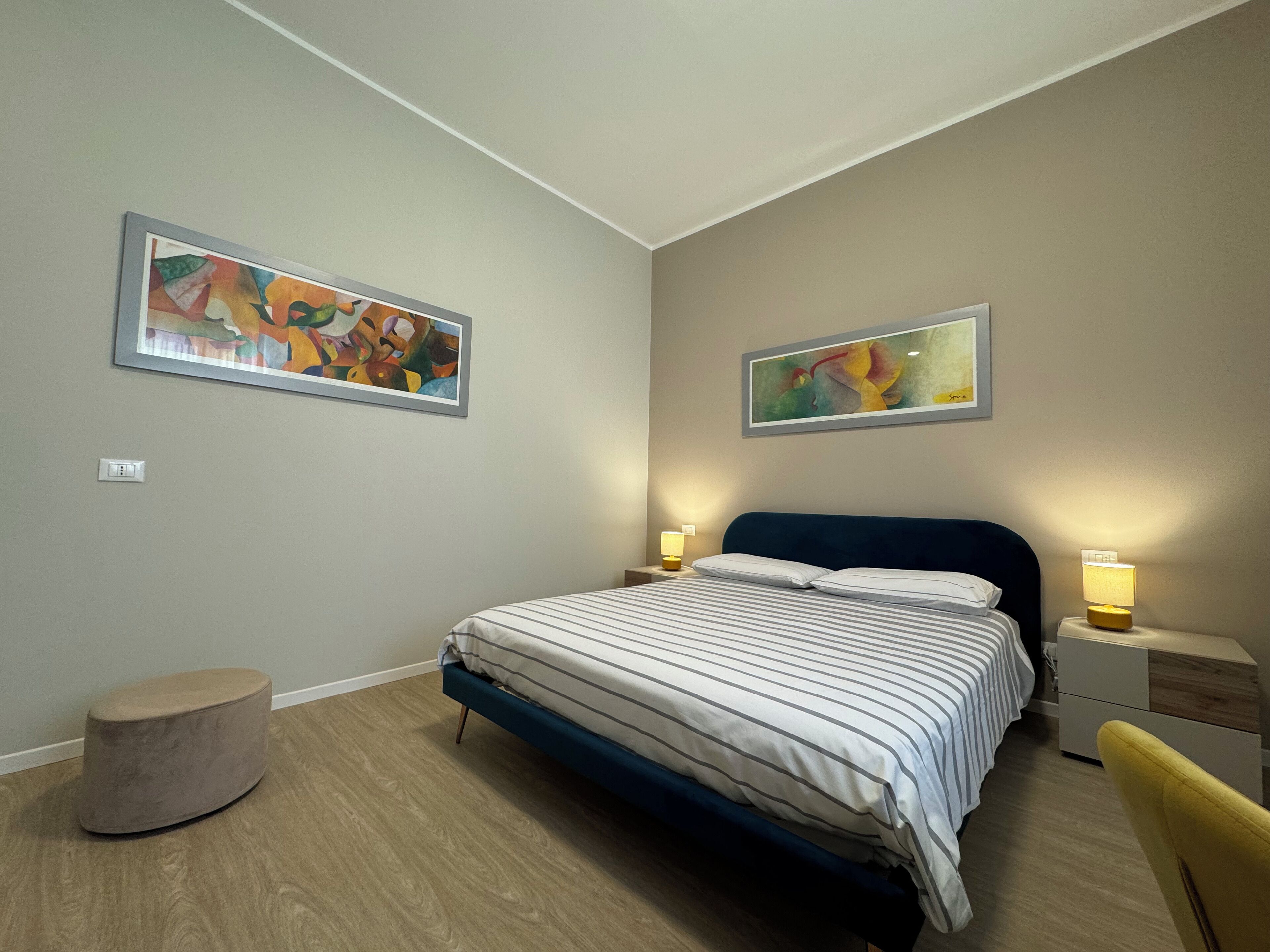 A modern bedroom with a bed, two bedside tables, a round ottoman, and two abstract paintings on the walls.