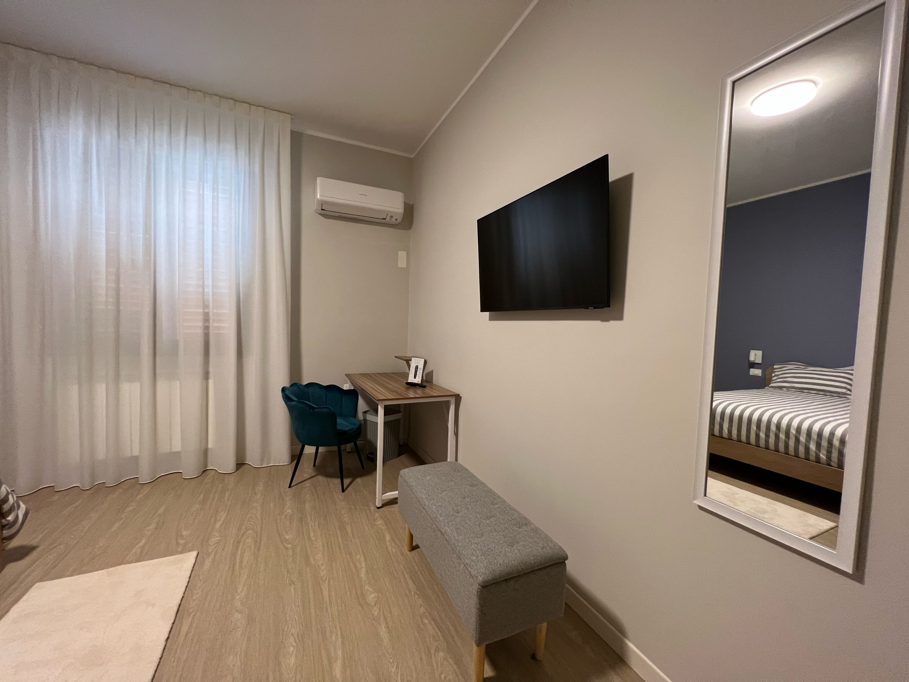 A compact hotel room with a wooden floor, a desk, a chair, a bench, a flat-screen TV mounted on the wall, and a large mirror reflecting the bed.