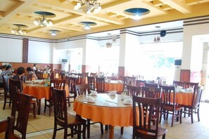 Un espace repas comprenant plusieurs tables et chaises, un plafond haut avec des poutres apparentes et de grandes fenêtres laissant entrer la lumière naturelle.