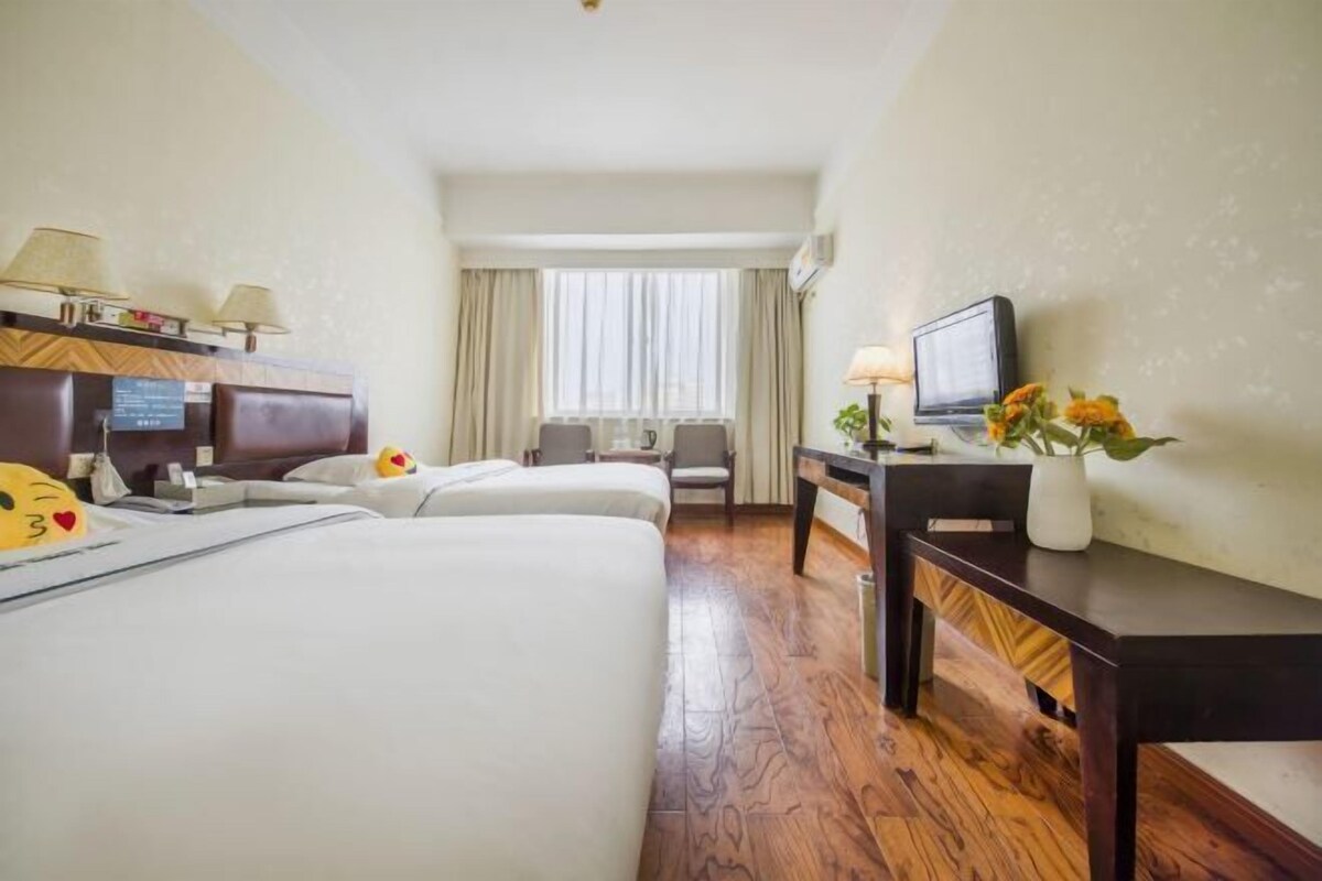 A hotel room with two beds, a desk with a TV, a window with curtains, and a wall-mounted TV.