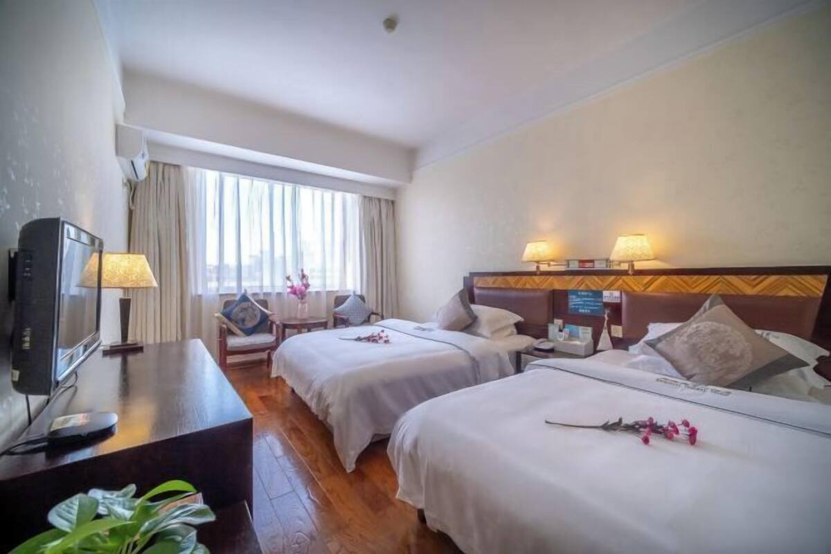 Ein Hotelzimmer mit zwei Betten, einem Schreibtisch mit Fernseher, einem Sessel und einem Fenster mit Vorhängen.