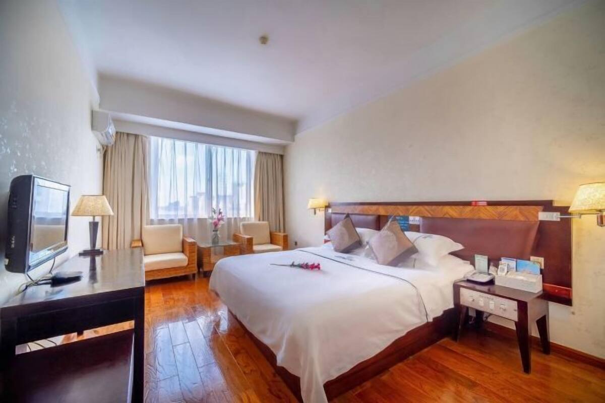 A hotel room with a bed, bedside tables, a TV, a window with curtains, and a wooden floor.