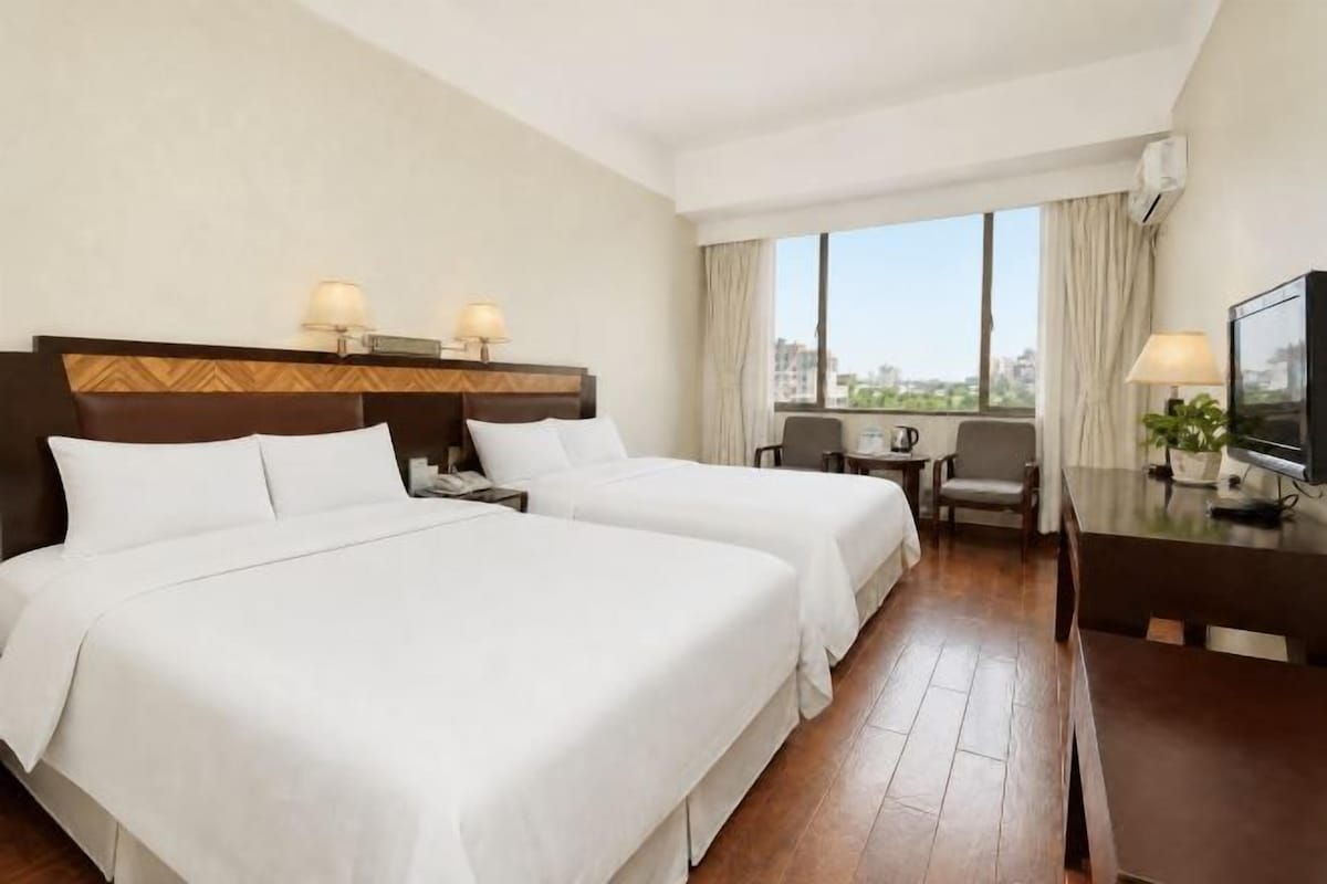 Habitació d'hotel amb dos llits, escriptori, cadira, televisió i una finestra gran amb vista a la ciutat.